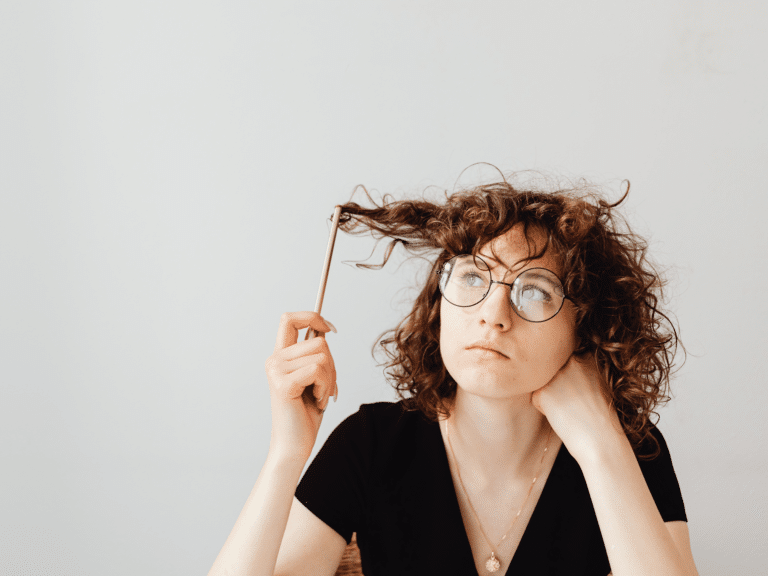 young woman wearing a black shirt with medium length orange hair wearing eye glasses. she is wrapping a pencil around the end of her curly hair