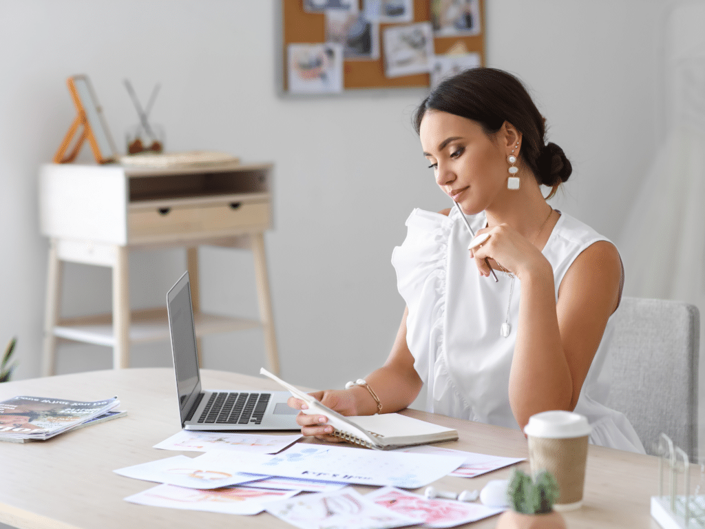 woman sitting at a table behind a computer reading something written on a notebook